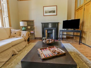 A living room with a white sofa, a backgammon board on a coffee table, a fireplace, a television on a stand, and a framed picture above the fireplace at The Residence in Cleator