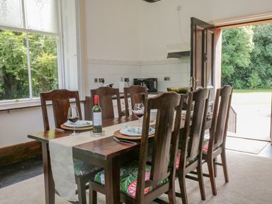 A dining room with a wooden table and chairs with floral seat cushions next to a large window and open door showing a garden at The Residence in Cleator