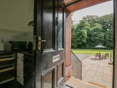 A black door partially open showing a kitchen with shelves and a microwave leading to an outdoor patio with a table umbrella and chairs at The Residence in Cleator