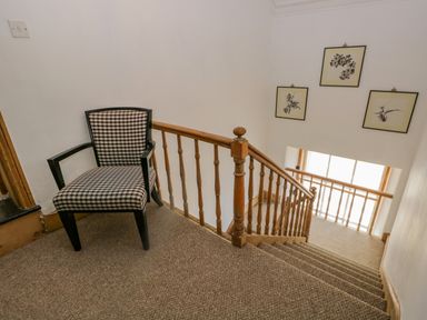 A staircase with wooden railing and a checkered chair on carpeted landing with framed pictures on the wall at The Residence in Cleator
