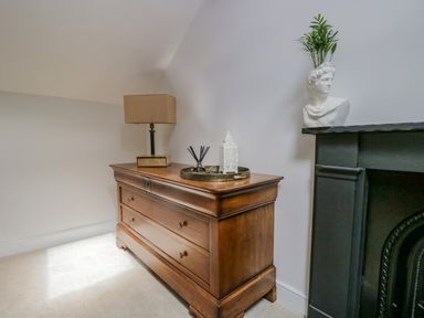 A wooden chest of drawers with a lamp, a tray with a diffuser, and a decorative lantern next to a black fireplace with a bust and a plant at The Residence in Cleator