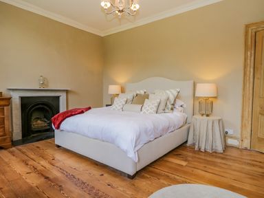 A bedroom with a bed with multiple pillows a side table with a lamp a fireplace and wooden floor at The Residence in Cleator