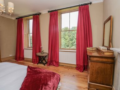 A bedroom with windows covered by red curtains a wooden dresser with a mirror and a small round table with a plant at The Residence in Cleator