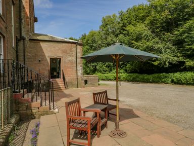 Two wooden chairs with a small table and green umbrella on a stone patio outside a brick building with trees in the background at The Residence in Cleator