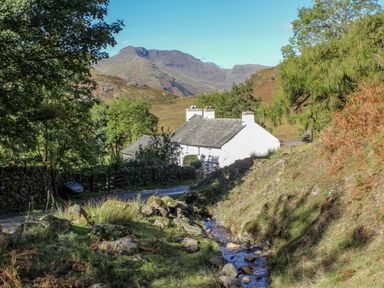 A small white house near a narrow stream with a mountain in the background at The Residence in Cleator