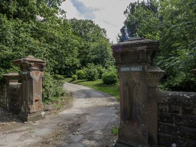 A stone gate entrance with pillars and a gravel driveway surrounded by trees and bushes at The Residence in Cleator