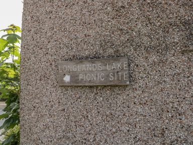 A wooden sign on a pebble textured wall pointing left for Longlands Lake picnic site at The Residence in Cleator