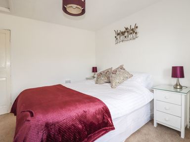 A bedroom with a bed covered with a red blanket and two patterned pillows with white nightstands and purple lamps at Gwynn Cottage in St Mabyn