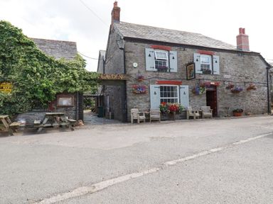 A stone building with wooden chairs and flower pots outside next to an entrance with a garden sign at Gwynn Cottage in St Mabyn