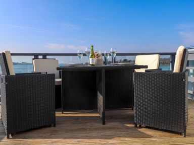 An outdoor dining table with chairs and wine glasses on a wooden deck overlooking water at White Sails in Y Felinheli