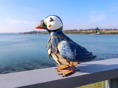 A metal puffin sculpture on a wooden railing overlooking a body of water with land in the background at White Sails in Y Felinheli