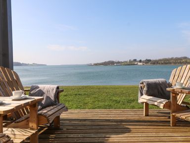 Two wooden chairs with throw blankets and cups on small tables on a wooden deck facing a body of water at White Sails in Y Felinheli