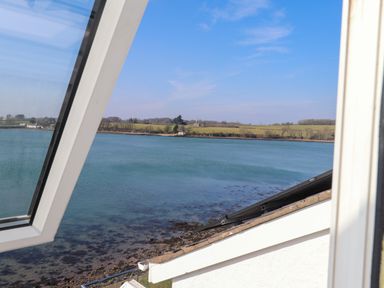 View of a body of water with shoreline and fields in the distance seen through an open window at White Sails in Y Felinheli