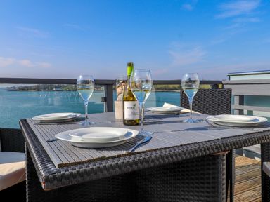 An outdoor dining table set with plates glasses and a bottle of wine overlooking a body of water at White Sails in Y Felinheli