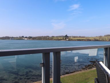 A view of a river with trees and houses on the opposite bank seen from a balcony with a glass railing and wicker chair at White Sails in Y Felinheli