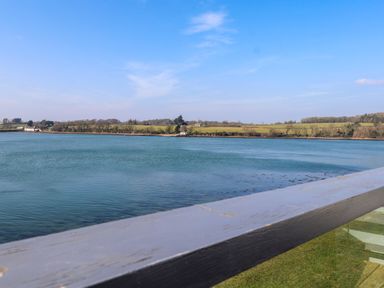 A body of water with grassy fields and trees in the background seen from behind a railing at White Sails in Y Felinheli