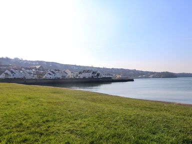 A grassy area beside calm water with houses along the shoreline and hills in the background at White Sails in Y Felinheli