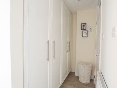 A hallway with white built-in wardrobes on the left wall a white wicker laundry basket on the floor and three framed pictures on the wall at White Sails in Y Felinheli