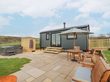 A patio with wooden furniture a parasol a hot tub and a small hut outside a corrugated metal cabin at Northcott Hut Stibb near Bude