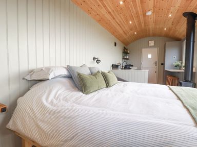 A bedroom with a bed with pillows a wooden ceiling a stove kitchen area and a door at Northcott Hut in Stibb near Bude