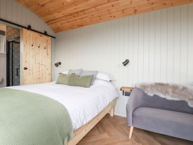 A bedroom with a wooden bed with green pillows and blanket a small gray sofa and a sliding wooden door at Northcott Hut Stibb near Bude