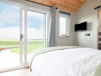 A bedroom with a bed facing a sliding glass door overlooking a grassy yard and a wall-mounted TV at Northcott Hut in Stibb near Bude