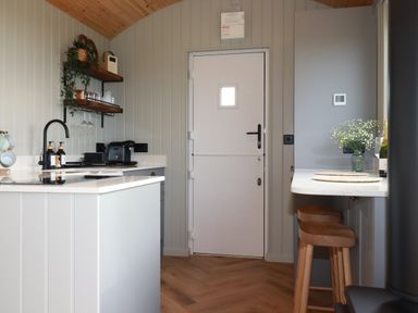 A kitchen with a white door a countertop with bar stools shelves with glasses and a faucet at Northcott Hut in Stibb near Bude