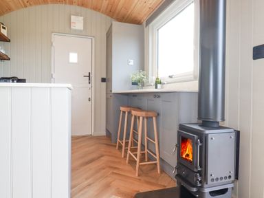 A kitchen area with a stove on the right two wooden stools under a counter and a window at Northcott Hut in Stibb near Bude