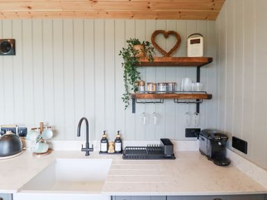 A kitchen sink with black faucet and soap dispensers next to kitchen counter with cups coffee machine and hanging glasses at Northcott Hut in Stibb near Bude