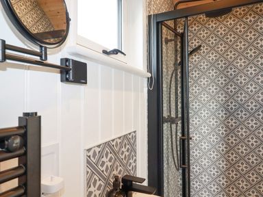 A bathroom with patterned tiled walls a black shower door a round mirror and a window at Northcott Hut in Stibb near Bude
