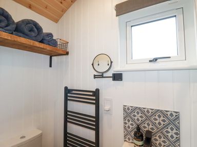 A bathroom with a towel rack a window a small round mirror a wooden shelf with towels and a sink with patterned tiles at Northcott Hut in Stibb near Bude
