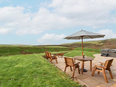 An outdoor patio with wooden chairs and table with umbrella and hot tub in a grassy area at Northcott Hut in Stibb near Bude
