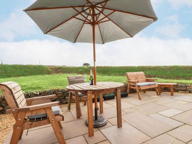 An outdoor patio with wooden chairs a round table with umbrella and a bottle on the table at Northcott Hut in Stibb near Bude