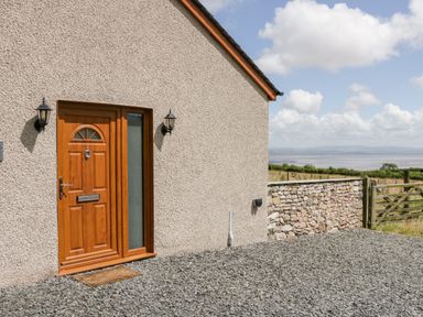 A wooden front door with side window lights on a pebble driveway beside a stone wall and countryside at Little Barn in Grange-Over-Sands near Cartmel