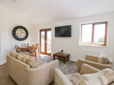 A living room with beige sofas a wooden dining table chairs a wall clock a wall-mounted TV a window and glass doors at Little Barn in Grange-Over-Sands near Cartmel
