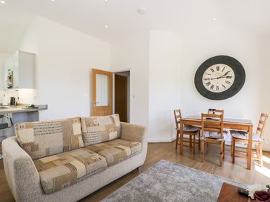 A living room with a sofa and a dining table with chairs and a large wall clock at Little Barn in Grange-Over-Sands near Cartmel