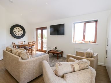 A living room with beige sofas a coffee table a wall-mounted TV a wooden dining table with chairs and a large clock on the wall at Little Barn in Grange-Over-Sands near Cartmel