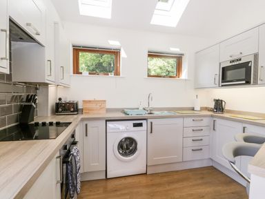 A kitchen with white cabinets wooden countertops a washing machine toaster microwave kettle and two small windows at Little Barn in Grange-Over-Sands near Cartmel