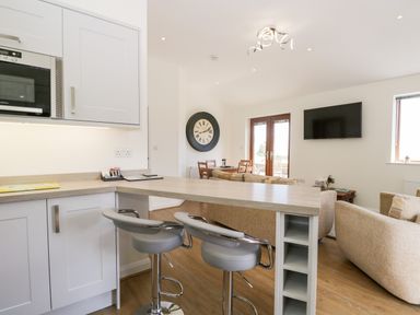 A kitchen area with two bar stools at a counter and a living room with sofas a dining table a clock and a TV mounted on the wall at Little Barn in Grange-Over-Sands near Cartmel