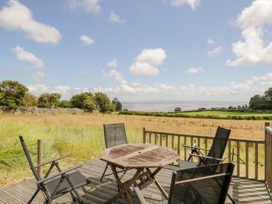 A wooden deck with a table and four chairs overlooking a field and distant water at Little Barn in Grange-Over-Sands near Cartmel