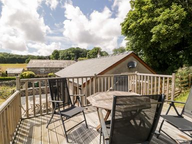 A wooden outdoor deck with a round table and four black chairs overlooking stone buildings and trees at Little Barn in Grange-Over-Sands near Cartmel