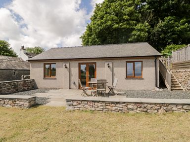 A single story building with stone walls and wooden doors and windows with an outdoor seating area at Little Barn in Grange-Over-Sands near Cartmel