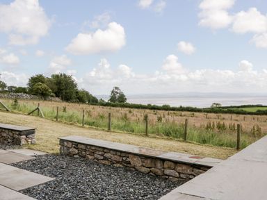 An outdoor patio with stone benches and gravel overlooking a fenced field and distant hills at Little Barn in Grange-Over-Sands near Cartmel