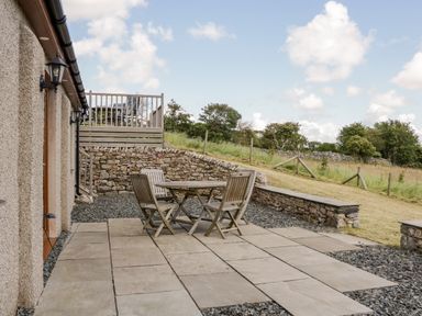 An outdoor patio with a round wooden table and three wooden chairs a stone wall and grass field at Little Barn in Grange-Over-Sands near Cartmel