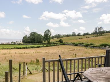 A wooden deck with a table and chairs overlooking grassy fields with trees and a stone wall at Little Barn in Grange-Over-Sands near Cartmel