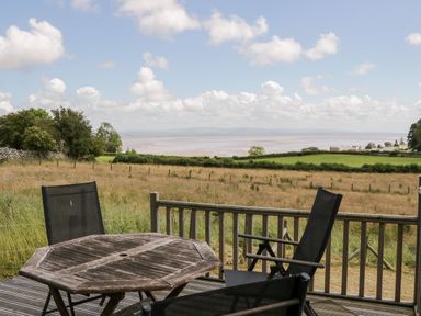 An outdoor deck with a wooden table and black chairs overlooking fields and water at Little Barn in Grange-Over-Sands near Cartmel