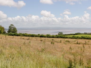 An open grassy field with a fence and hedgerows overlooking a body of water under a partly cloudy sky at Little Barn in Grange-Over-Sands near Cartmel