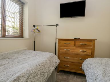 A bedroom with two beds covered in patterned bedding a wooden dresser with drawers a TV mounted on the wall and a window with blinds at Little Barn in Grange-Over-Sands near Cartmel
