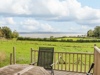 A wooden deck with outdoor chairs overlooking a grassy field and a large body of water at Little Barn in Grange-Over-Sands near Cartmel
