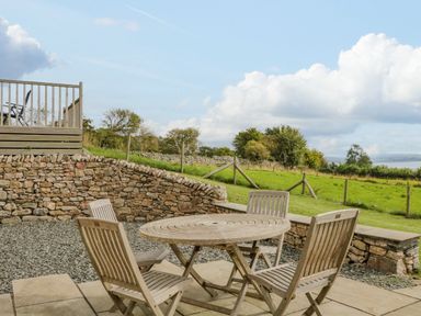 An outdoor patio area with a round wooden table and four wooden chairs near a stone wall overlooking a grassy field at Little Barn in Grange-Over-Sands near Cartmel